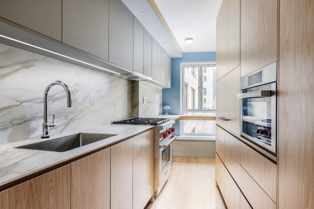 Modern galley kitchen with light wood cabinetry, marble backsplash, stainless steel sink and appliances, gas cooktop, and a bright window nook with natural light.