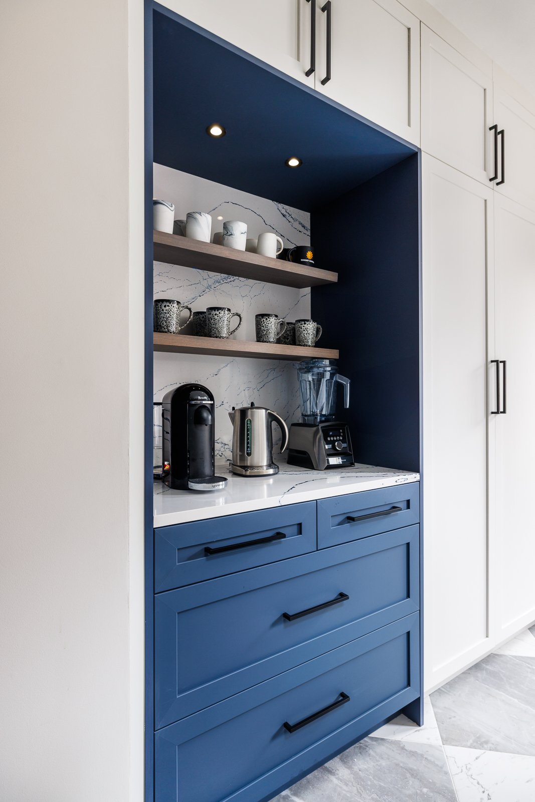 Compact kitchen beverage station with a bold navy blue built-in featuring shaker-style drawers, black hardware, and open wood shelving. The space includes a marble countertop with a coffee maker, kettle, and mugs neatly arranged for easy access. Surrounded by white cabinetry, the contrasting color scheme creates a modern and functional focal point in the kitchen.