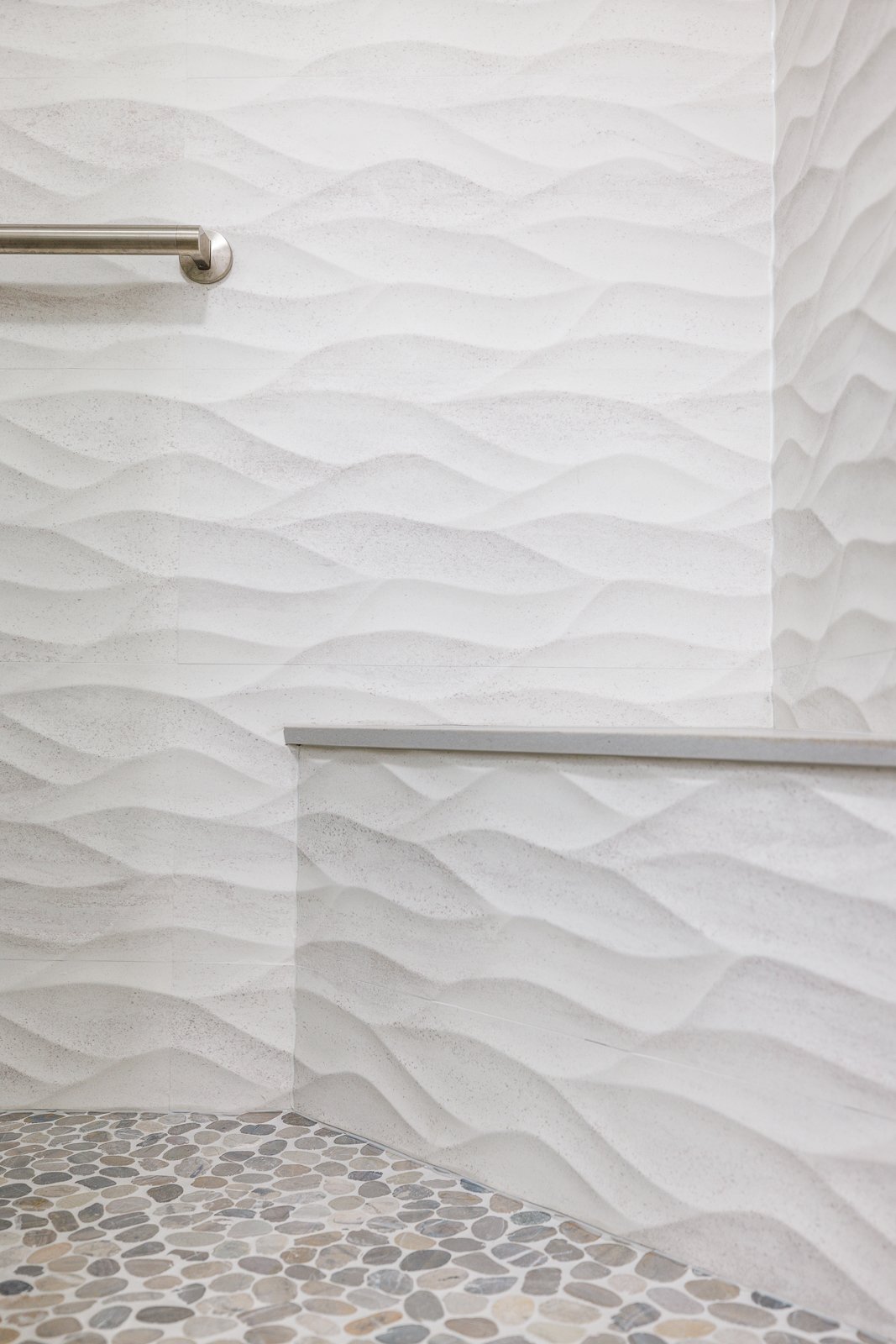 Close-up of a modern shower with textured white wave-patterned tiles, a built-in bench, stainless steel grab bar, and pebble-style stone flooring.