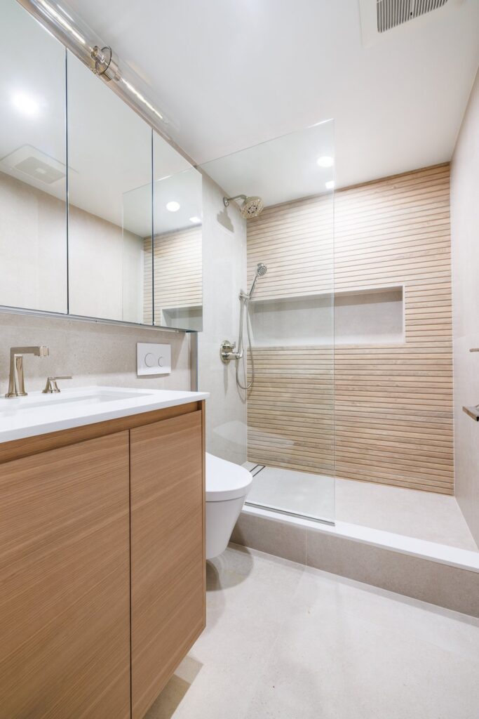 Home remodeling contractor on the West Side. Modern bathroom with light wood vanity, minimalist fixtures, wall-mounted toilet, and a walk-in shower featuring horizontal wood-textured tile and glass enclosure.