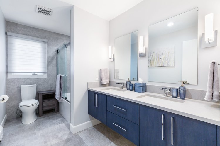 Contemporary bathroom with a double vanity featuring blue cabinets, dual mirrors with wall sconces, a toilet, and a glass-enclosed shower with a gray tile surround.