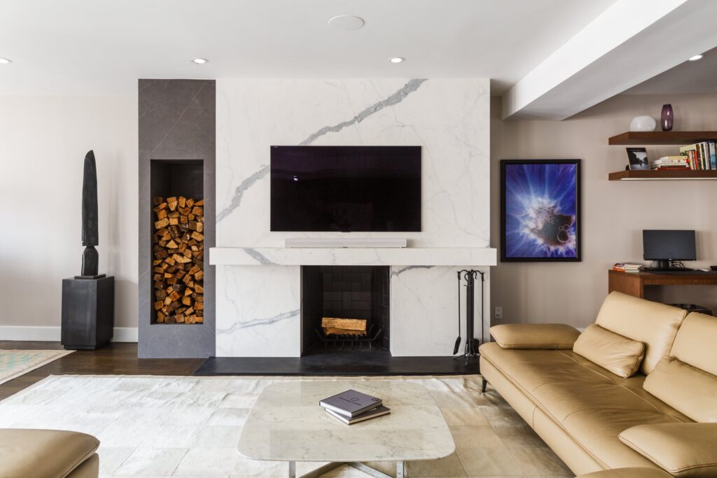 Contemporary living room with a marble fireplace wall, mounted flat-screen TV, stacked firewood niche, tan leather sectional sofa, and a modern coffee table on a neutral rug.