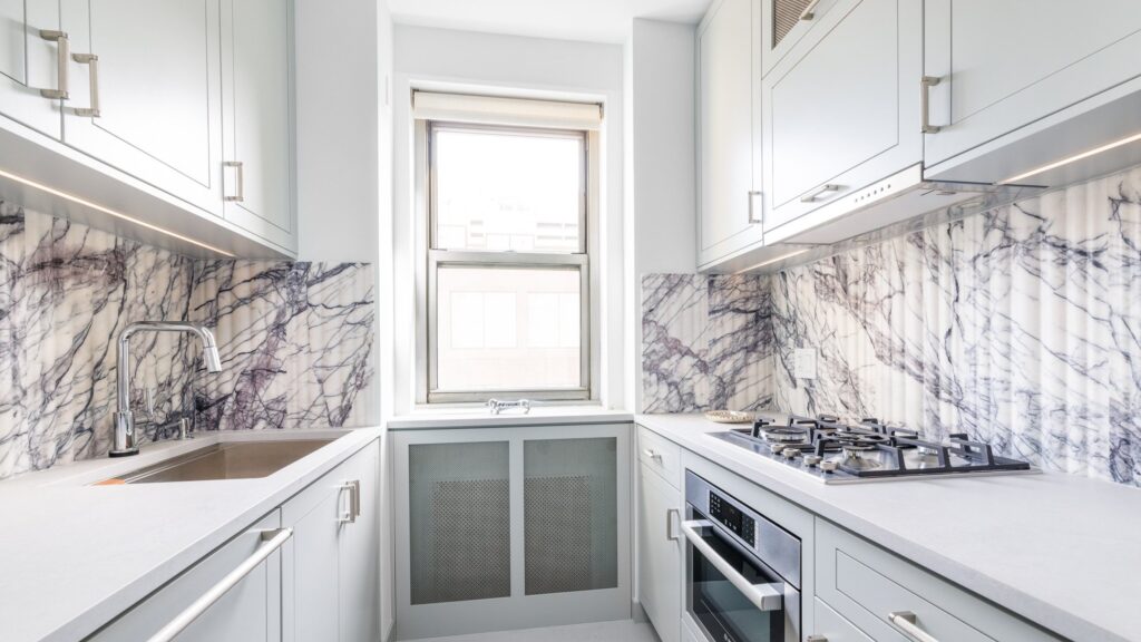 Bright galley kitchen with light gray cabinetry, marble-patterned backsplash, built-in gas cooktop and oven, under-cabinet lighting, and a large central window.