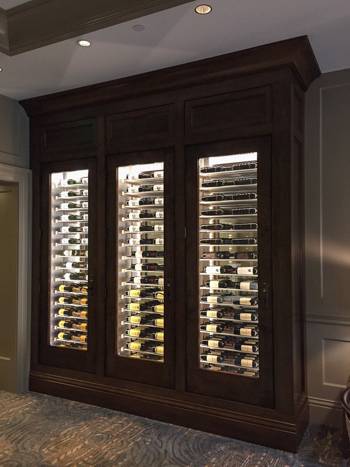 Elegant built-in wooden wine cabinet with three glass doors and interior lighting, showcasing multiple rows of horizontally stored wine bottles.