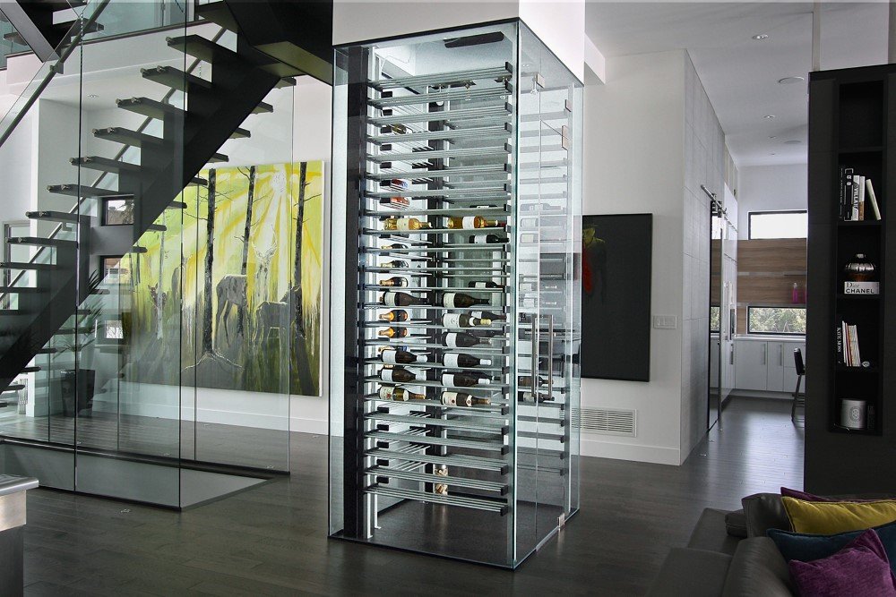 Contemporary living space featuring a glass-enclosed wine tower with horizontal metal racks, adjacent to a modern floating staircase and large abstract forest artwork.