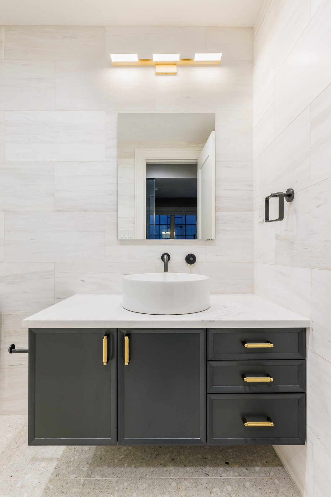 Modern bathroom vanity with a round vessel sink, matte black fixtures, dark cabinetry with gold hardware, and a white marble countertop against light tiled walls.