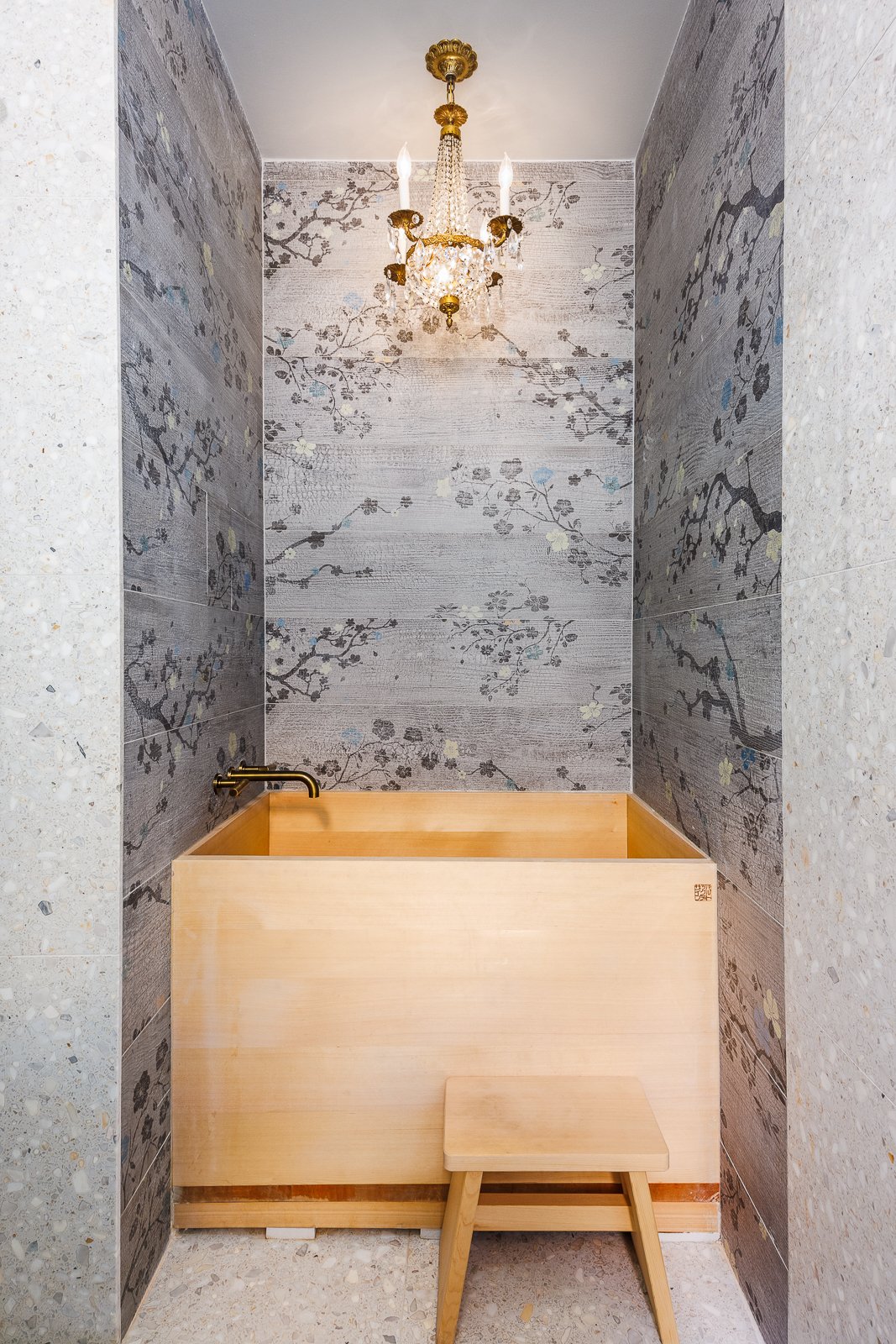 Serene bathroom featuring a light wood Japanese soaking tub, a small matching stool, floral-patterned wall tiles, and a decorative chandelier overhead.