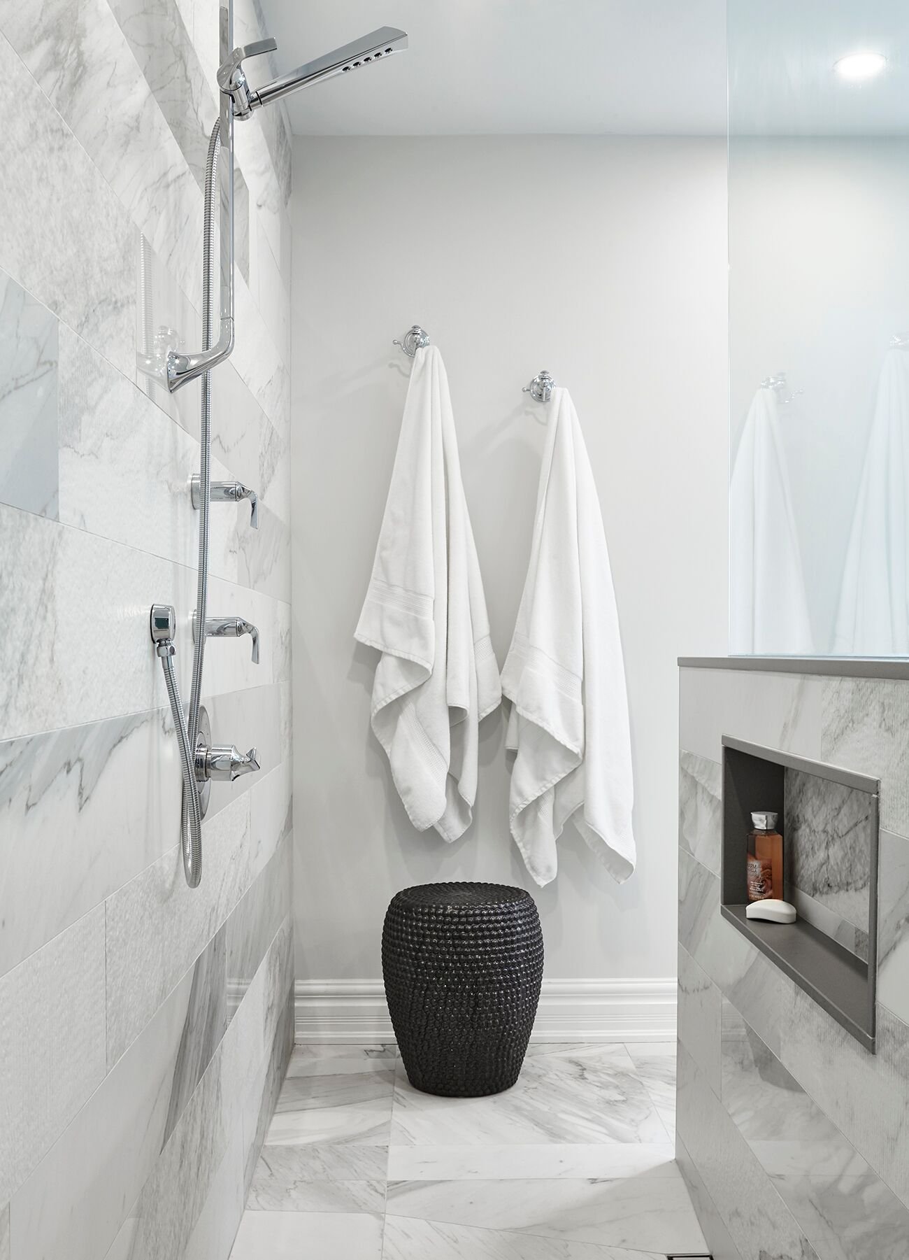 Stylish bathroom with marble tile walls and floors, a modern shower system with handheld and wall-mounted fixtures, two white towels hanging on the wall, a black woven stool, and a built-in niche holding toiletries.