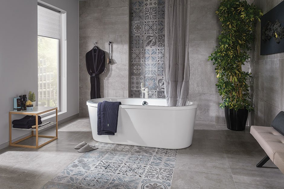 Contemporary bathroom with a freestanding white bathtub, patterned tile inlay, gray stone walls and floors, a robe hanging on the wall, and a tall indoor plant adding greenery.