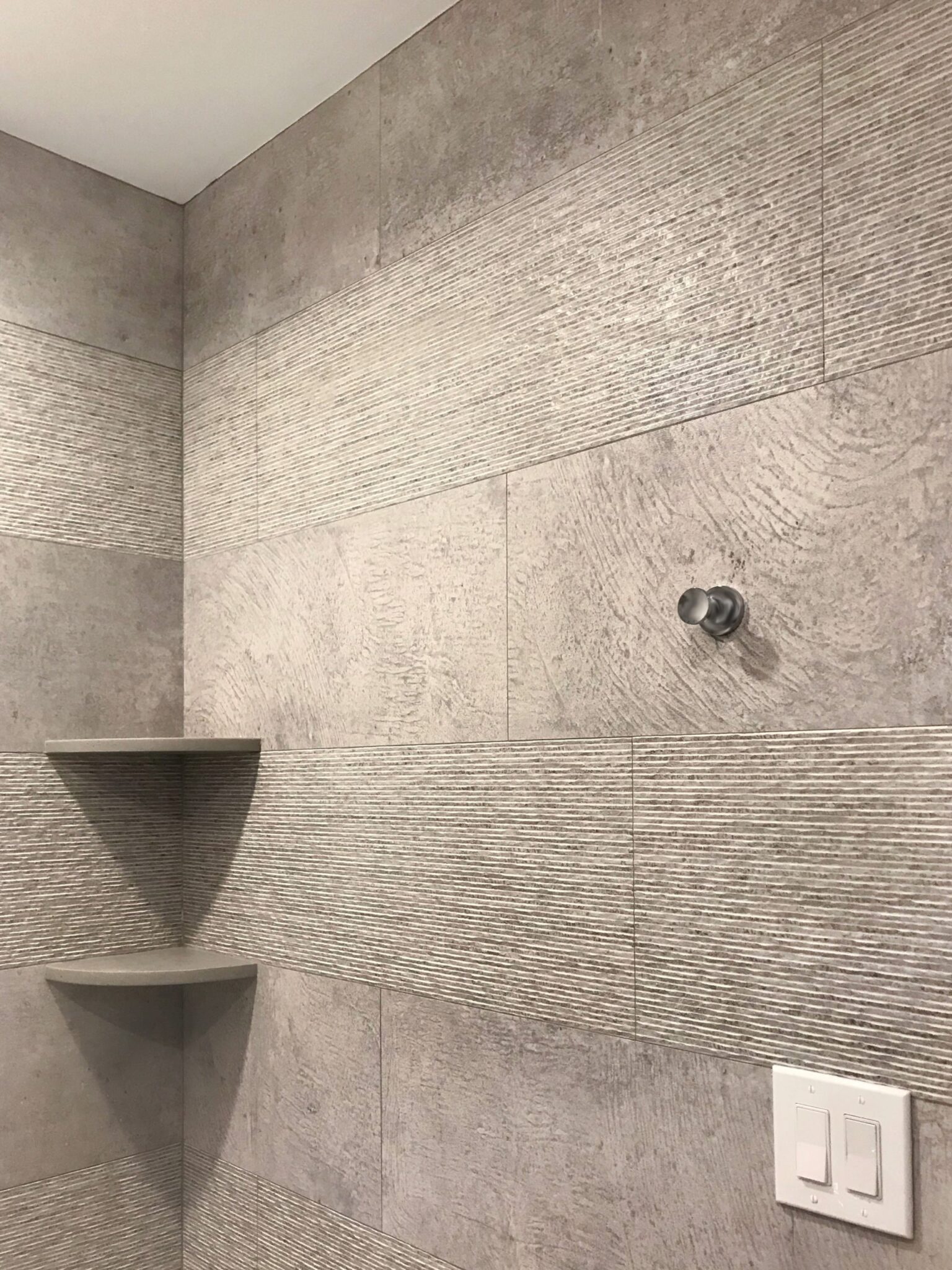 Modern shower wall with large textured gray tiles, built-in corner shelves, a metal fixture for a showerhead, and a nearby light switch.