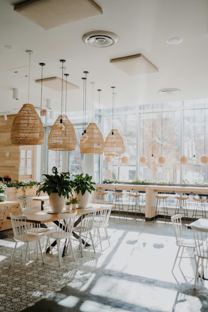 Bright and airy café interior with large floor-to-ceiling windows, white wooden chairs, light wood tables, woven pendant lights, indoor plants, and patterned tile flooring.