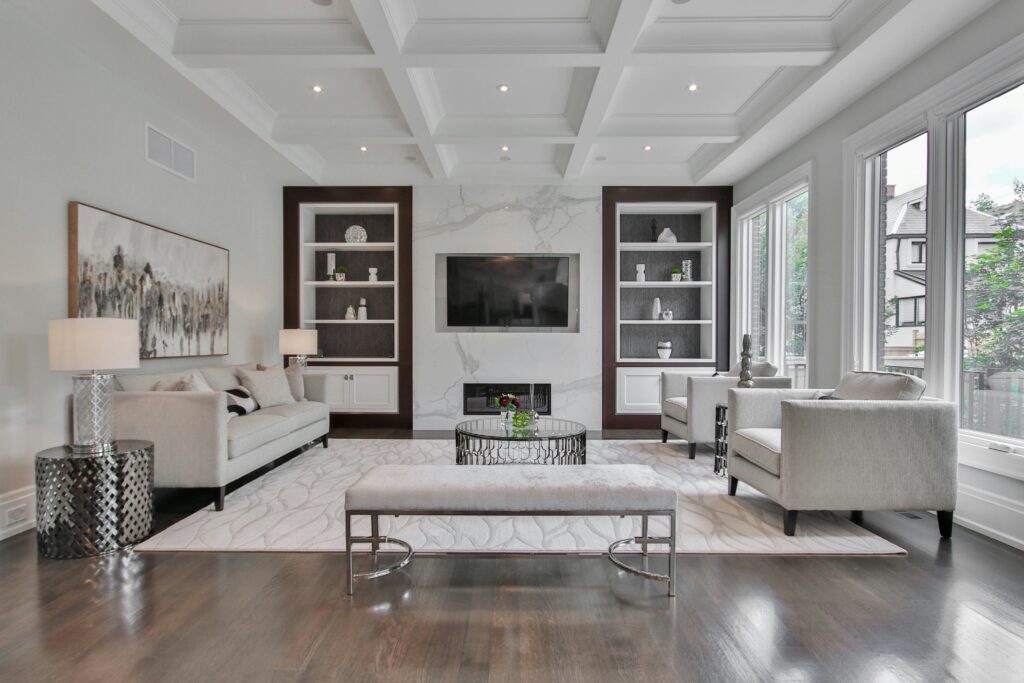 Elegant living room with coffered ceiling, marble fireplace wall, built-in shelving units, neutral-toned furniture, a large rug, and floor-to-ceiling windows providing natural light.