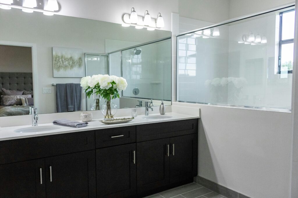 Home remodeling contractor in the Upper West Side. Modern bathroom with dark wood vanity, white countertop, dual sinks, a large mirror, decorative lighting, a glass-enclosed shower, and a vase of white flowers for decor.