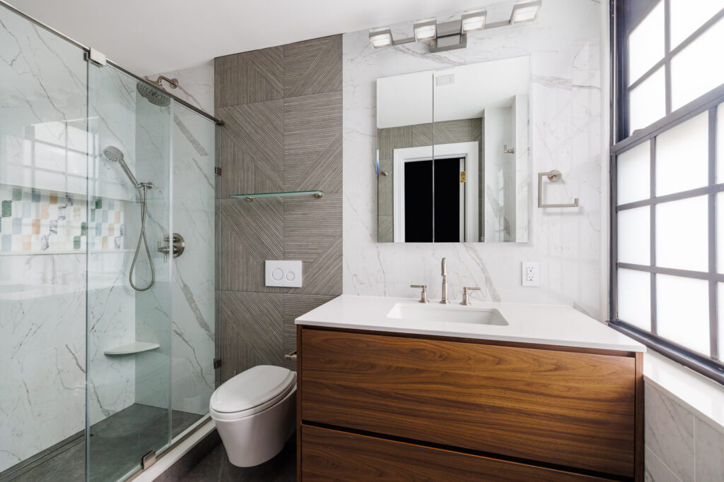 Contemporary bathroom with a light wood floating vanity, white countertop, and frameless mirror. The design features subtle beige walls, a glass shower enclosure, and a minimalist approach to maximize small space functionality.