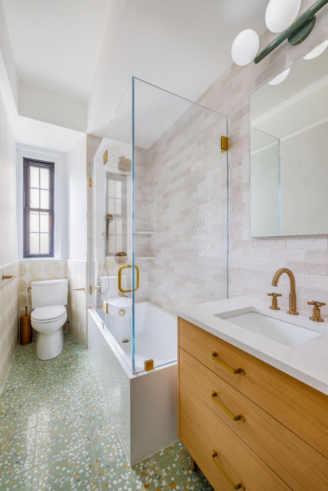 Elegant bathroom featuring a terrazzo-style green floor with multicolored chips, a white soaking tub with frameless glass enclosure and brass hardware, and a warm wood vanity topped with a white quartz sink. The space is finished with cream-toned zellige-style shower tiles, a globe light fixture, and a large window for natural light.