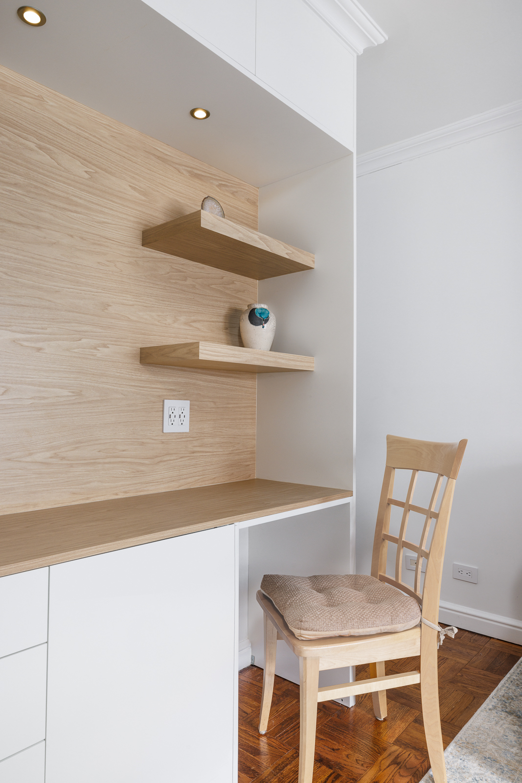 Minimalist wooden desk area with two floating shelves, a small ceramic vase, built-in outlet, overhead light, and cushioned wooden chair.