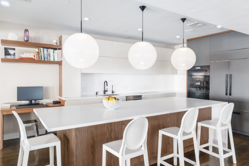 Modern, minimalist kitchen featuring a large white waterfall island with wood paneling and four sleek white chairs. Above the island hang three large globe pendant lights. The back wall includes handle-less white cabinetry, a matte black faucet and sink, and integrated appliances within dark grey cabinetry. A built-in desk with a computer, books, and floating shelves adds functionality to the left side of the space. The overall aesthetic is bright, clean, and contemporary.