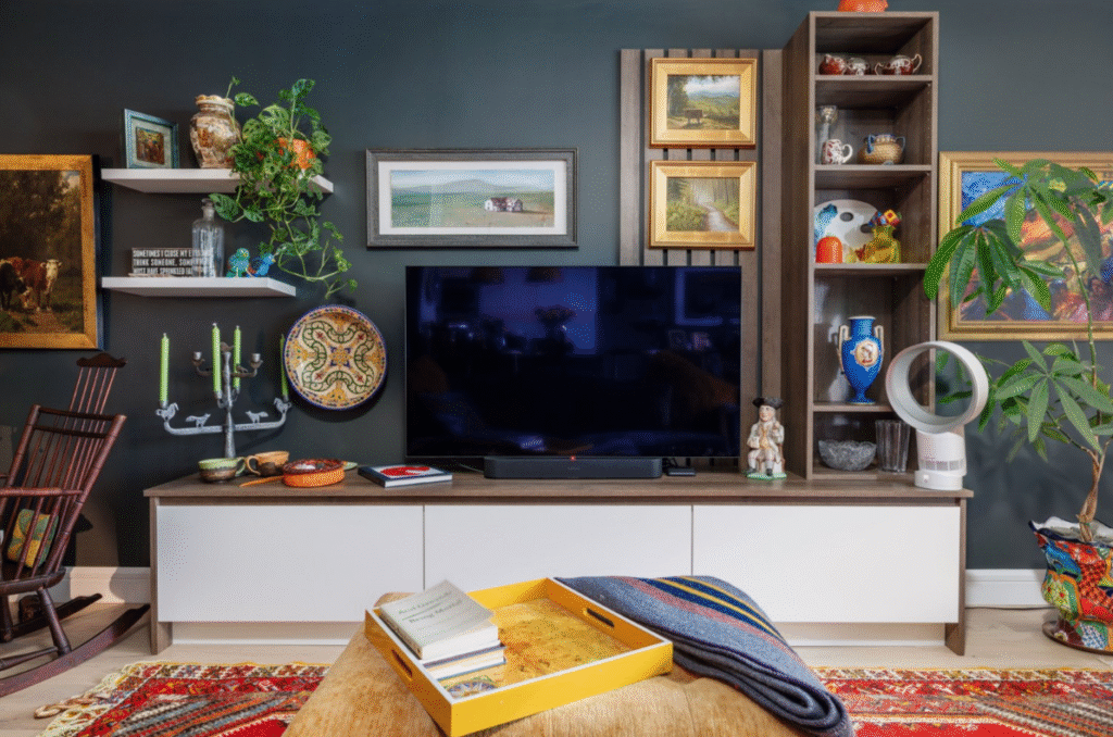 Eclectic and cozy living room featuring a dark accent wall with a mounted flat-screen TV surrounded by decorative art and collectibles. The TV console includes a mix of white cabinetry and wood accents, with open shelves displaying vintage ceramics, framed artwork, a candelabra, potted plants, and a bladeless fan. A vibrant red patterned rug adds warmth beneath a wooden coffee table topped with a yellow tray, books, and a folded striped blanket. The overall space blends modern tech with classic charm and curated personality.