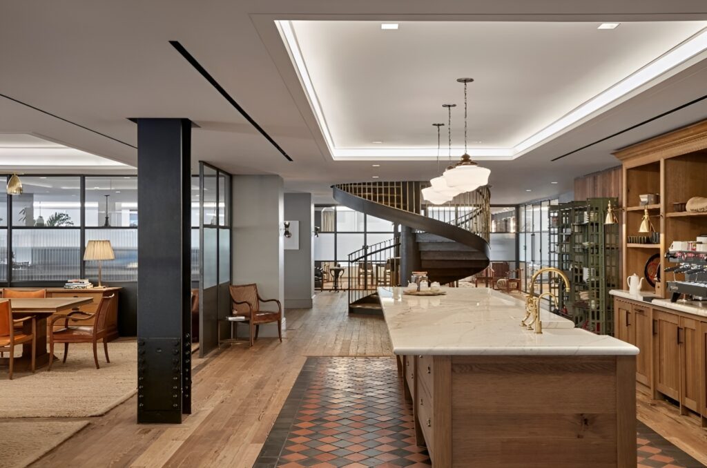 Elegant open-concept interior featuring a spacious kitchen with a large marble island, brass fixtures, and wood cabinetry. A patterned tile inlay runs beneath the island, contrasting with the surrounding hardwood floors. To the right, a coffee station and open shelving display curated kitchenware. Overhead, pendant lights hang from a recessed coffered ceiling with ambient lighting. A dramatic black spiral staircase with brass railings anchors the back of the room, leading to an upper level. On the left, glass-partitioned offices and a conference area with leather chairs and a table offer a warm, refined workspace aesthetic.