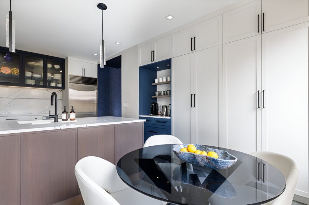 Modern Upper East Side kitchen with a sleek design featuring white shaker cabinets, a marble backsplash, and a wood-paneled island with a waterfall countertop. The space includes black pendant lighting, a built-in stainless steel refrigerator, and a round glass dining table with white upholstered chairs. A decorative blue bowl filled with lemons adds a pop of color against the minimalist neutral tones.