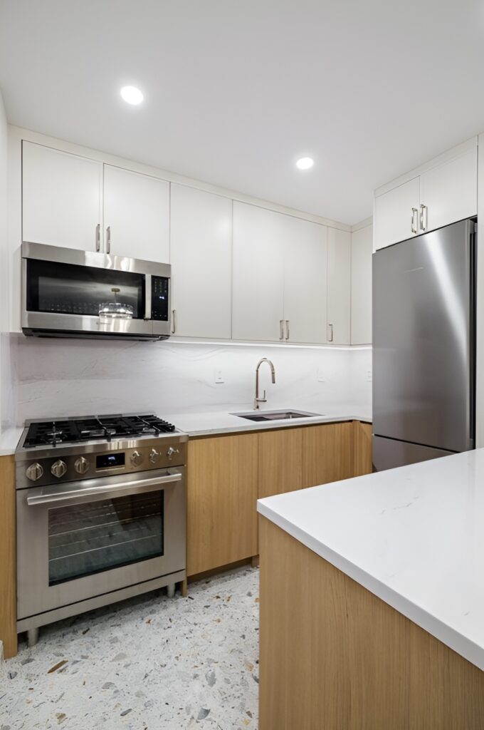 Contemporary Midtown East kitchen with a clean, minimalist design featuring white upper cabinets and natural wood lower cabinetry. The space includes stainless steel appliances, such as a gas range, built-in microwave, and refrigerator, paired with white quartz countertops and a subtle marble backsplash. The terrazzo-style flooring adds texture and visual interest to the compact yet functional layout.