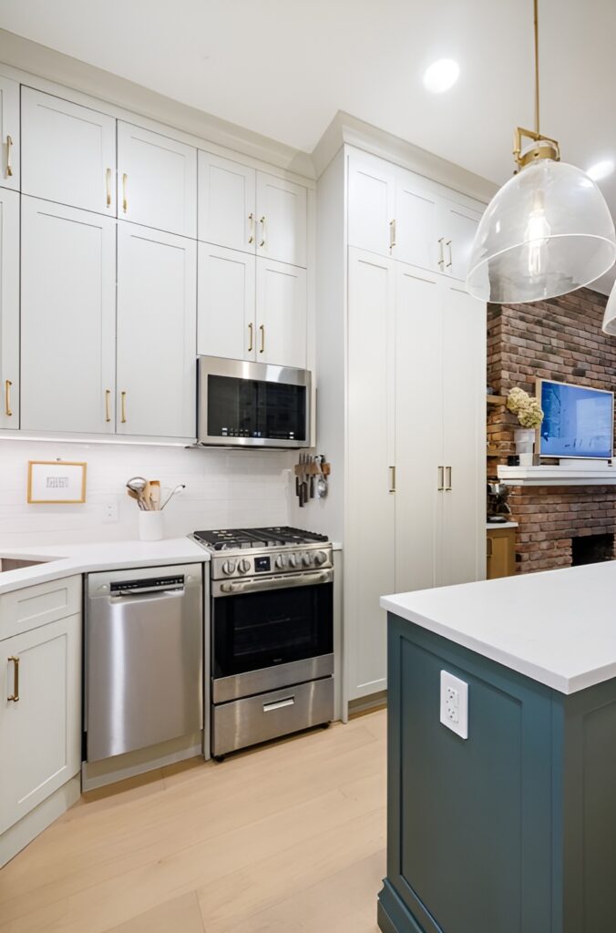 Charming West Side kitchen with tall white shaker cabinets accented by brass hardware, paired with stainless steel appliances, including a built-in microwave, dishwasher, and gas range. The space features a white subway tile backsplash, light wood flooring, and a large island with a muted teal base and white quartz countertop. An exposed brick wall with a mounted TV adds character and warmth to the open layout. Brownstone renovation in Manhattan.