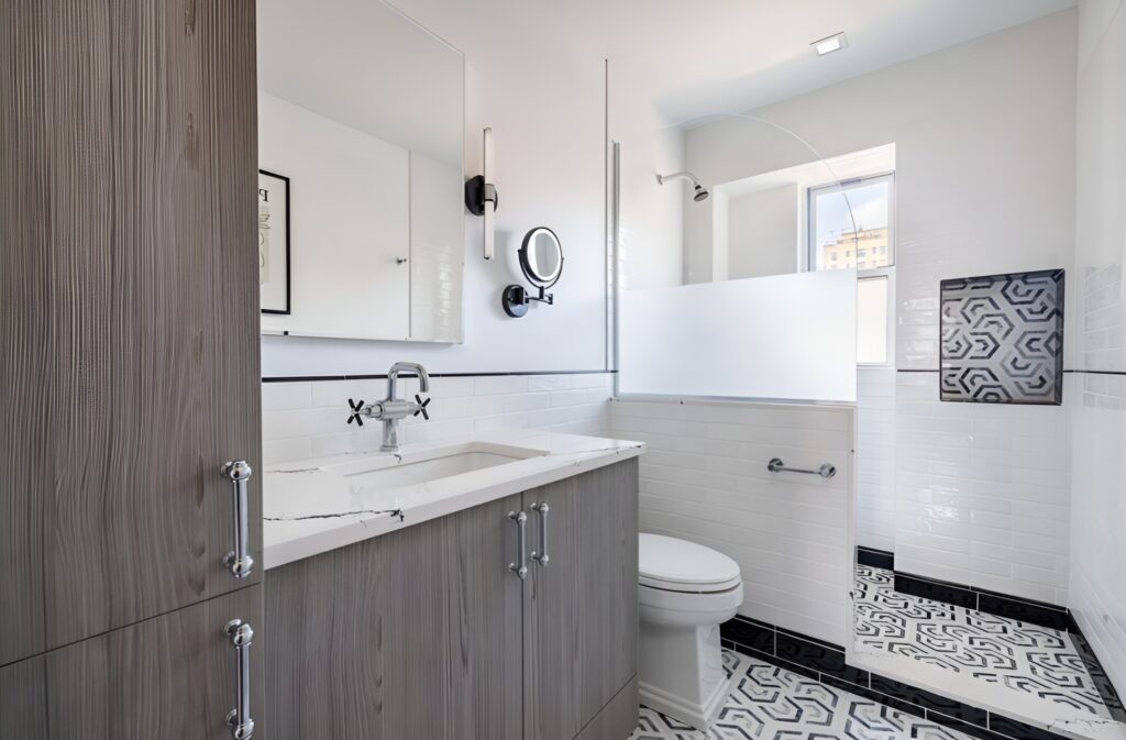 Stylish Upper West Side bathroom with a modern design featuring white subway tile walls, a floating vanity in a gray wood finish, and a marble-look countertop. The walk-in shower includes a frosted glass divider and geometric patterned tile accents that continue onto the floor. Black fixtures and hardware add a bold contrast to the otherwise neutral palette.