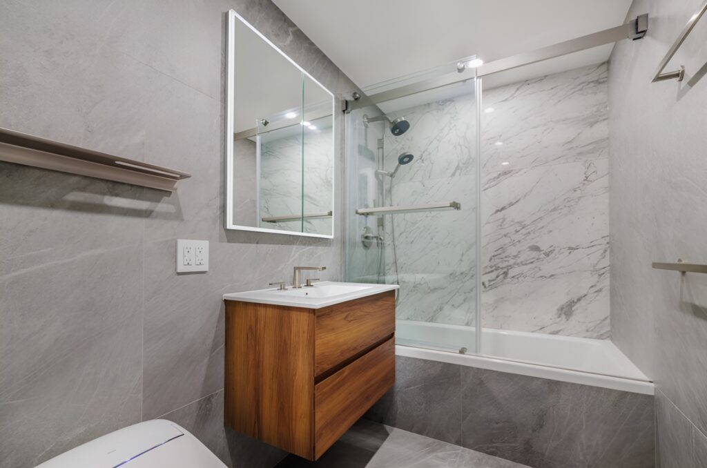 Contemporary Lincoln Square bathroom with a sleek design featuring gray stone-look wall tiles and a white bathtub with a marble-style surround. The vanity has a floating wooden cabinet with clean lines and a white countertop, paired with a frameless mirror. A glass sliding shower door and modern chrome fixtures complete the minimalist aesthetic.