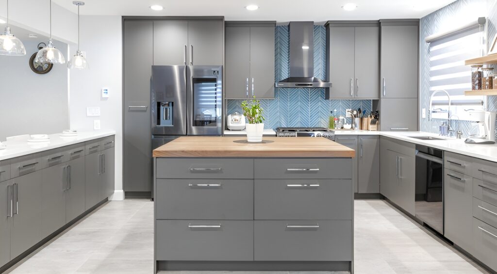 Spacious Yorkville kitchen featuring sleek gray cabinetry with chrome handles and a matching central island topped with a warm butcher block surface. A striking blue chevron backsplash adds texture behind the stainless steel range hood and stove. The design includes modern pendant lighting, integrated appliances, and a mix of open shelving and closed storage for a contemporary yet functional layout.