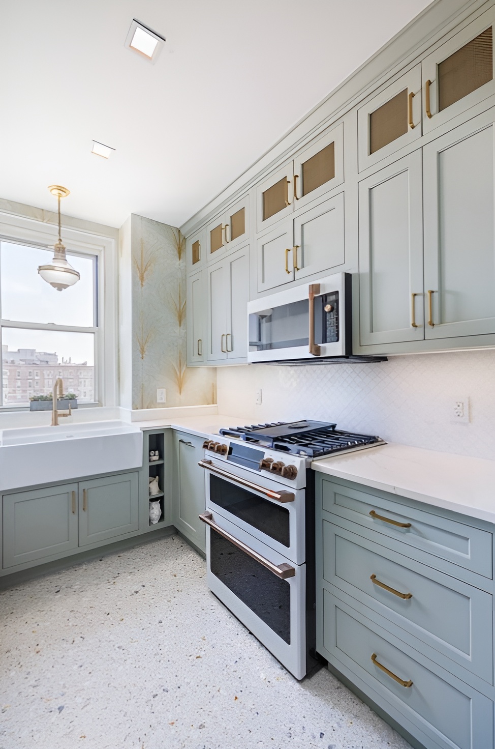 Elegant kitchen with pastel sage green cabinetry accented by brass hardware and mesh panel inserts. The design features a farmhouse sink beneath a large window, a white quartz countertop, and a modern dual-fuel range with a matching built-in microwave. Patterned wallpaper, terrazzo-style flooring, and a brass pendant light add vintage charm to the bright and airy space.