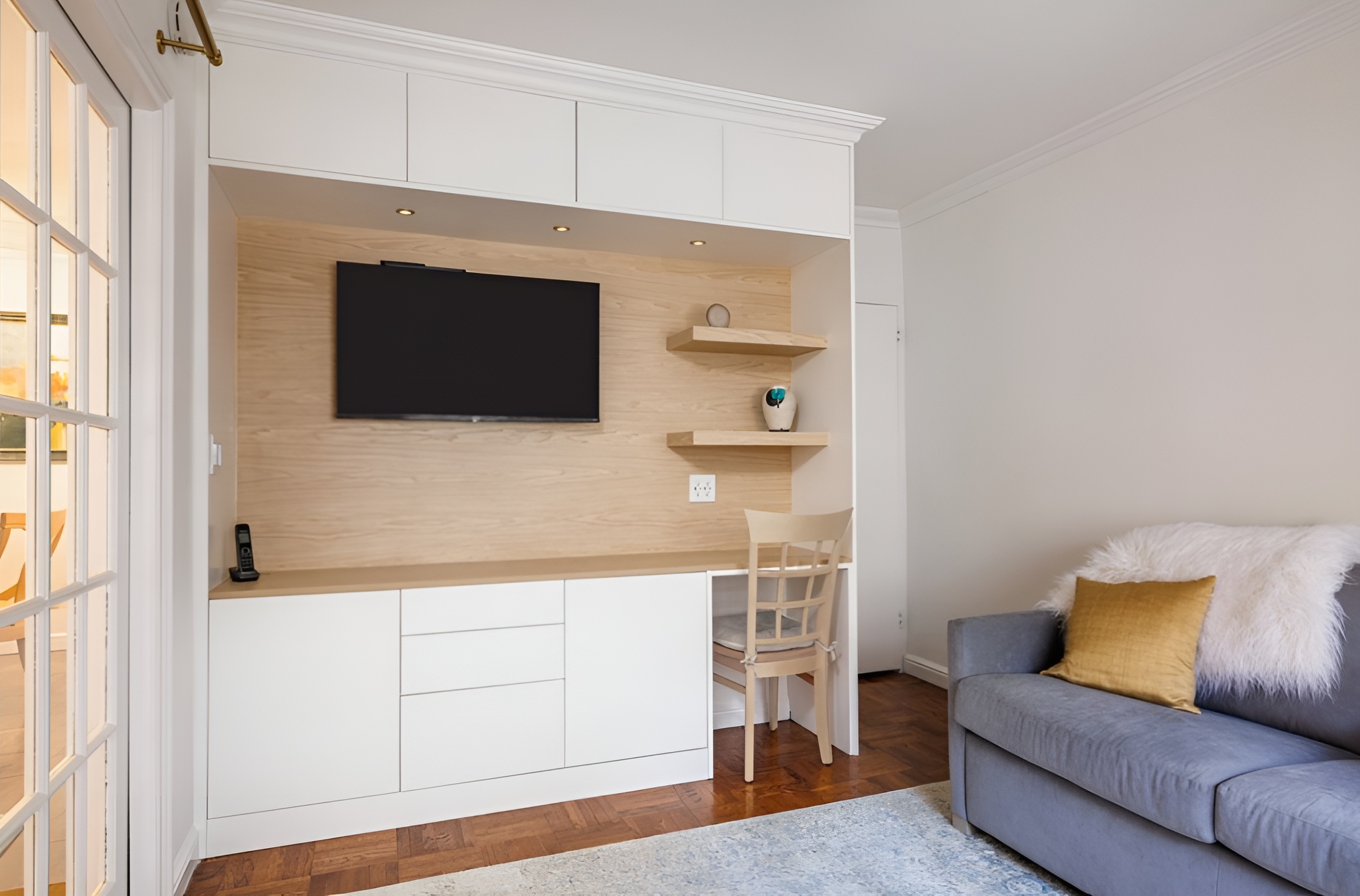 Compact living area featuring a built-in wall unit with white cabinetry and light wood accents, combining a mounted TV, floating shelves, and a small desk workspace. A soft gray sofa with a faux fur throw and mustard pillow adds comfort, while French doors lead to an adjacent room, creating an open and airy feel with multifunctional design.