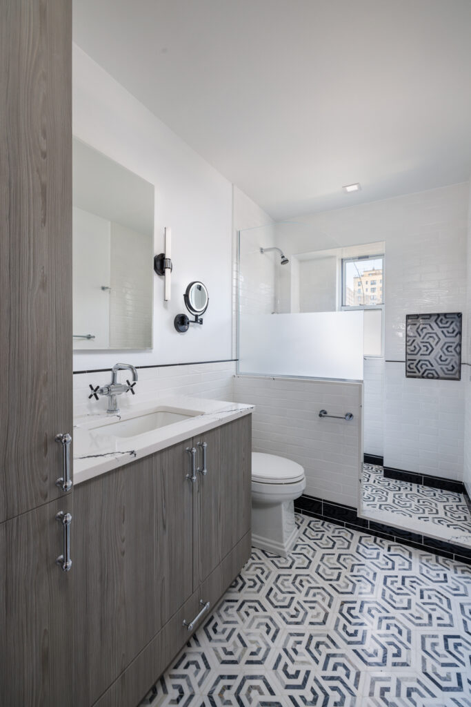 Bathroom with a gray wood vanity and marble countertop, white subway tile walls, a bathtub with a frosted glass panel, wall-mounted lighting, and a window providing natural light.