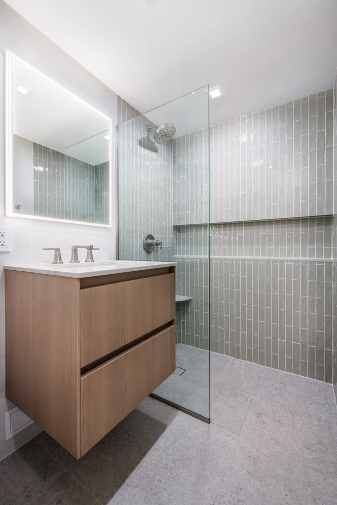 Modern bathroom with a wood vanity and white countertop, a glass-enclosed shower featuring vertical light gray subway tiles, built-in corner shelves, and brushed metal fixtures.