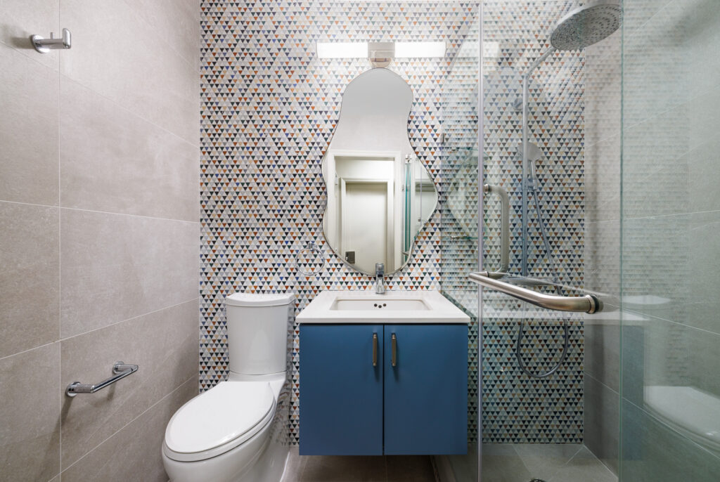 Bathroom with beige tiled walls, a patterned accent wall with geometric tiles, a unique irregular-shaped mirror, a blue vanity with a white countertop, a toilet, and a glass-enclosed shower with a metal grab bar.
