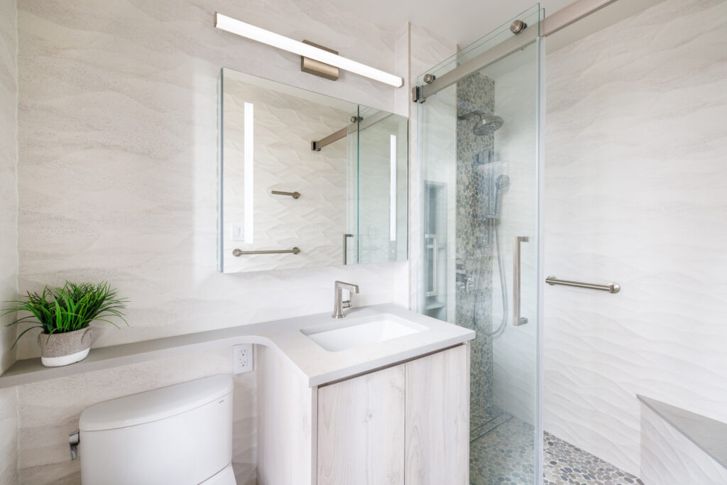 Bright modern bathroom with textured light beige walls, a wood vanity with a white countertop, a frameless mirror with built-in lighting, a glass-enclosed shower featuring a vertical mosaic accent strip, and a small potted plant on the counter.