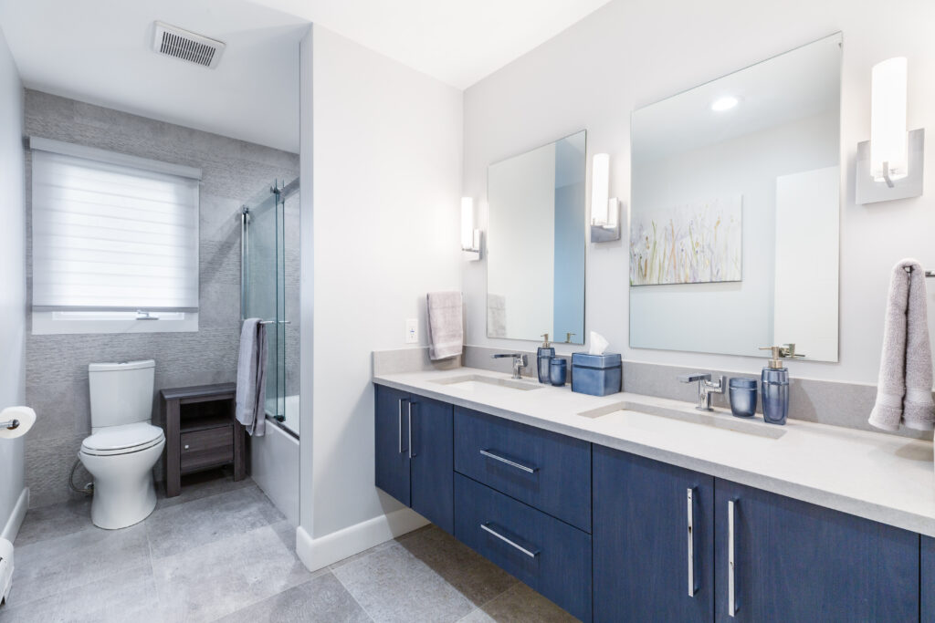 Contemporary bathroom with a double vanity featuring blue cabinets and white countertops, dual mirrors with modern wall lighting, a toilet next to a walk-in shower with glass doors, and a gray tile accent wall.
