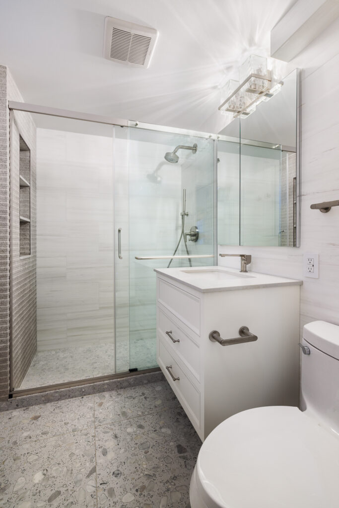 Modern bathroom with white marble-style walls, a glass-enclosed shower with a metal grab bar, a white vanity with a gray countertop, and built-in shelving with textured tile accents.
