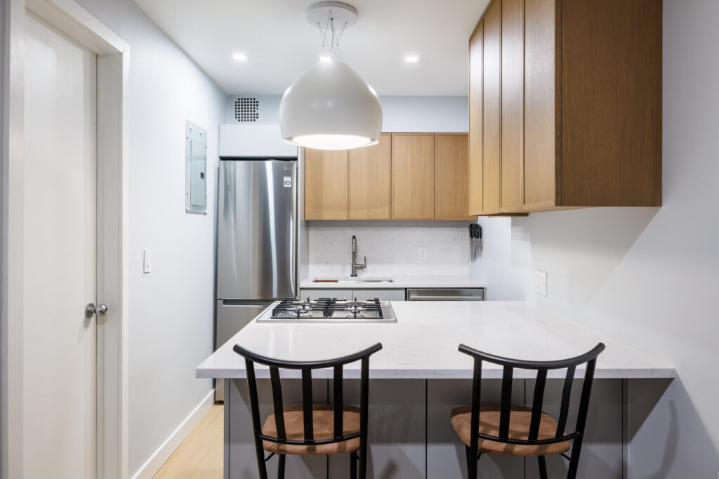 Compact kitchen with wood cabinetry, stainless steel appliances, white countertops, a gas cooktop on the peninsula, and two black bar stools