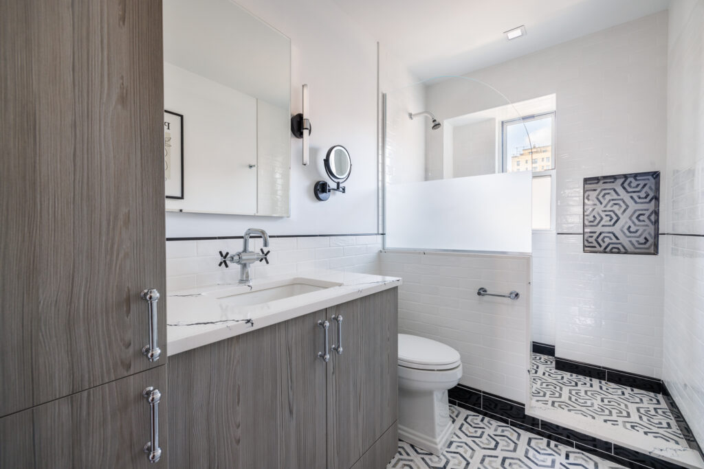 Modern bathroom with wood-textured vanity, white countertop, wall-mounted faucet, glass shower partition, and black-and-white patterned tile floor.