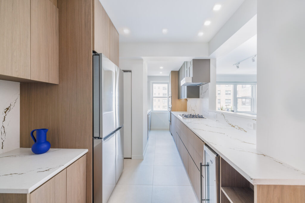 Modern galley kitchen with light wood cabinetry, marble countertops with gold veining, stainless steel appliances, and a blue decorative vase