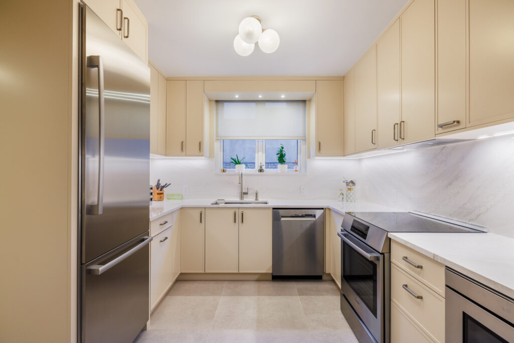 Contemporary kitchen with beige cabinetry, white marble countertops and backsplash, stainless steel appliances, and a window above the sink with plants