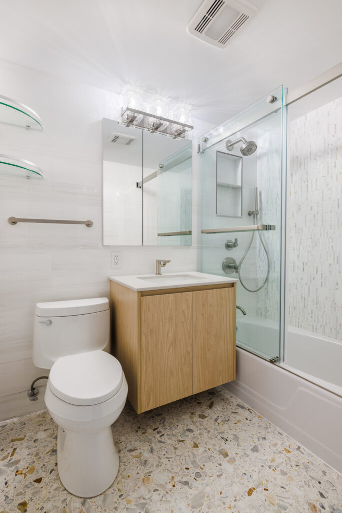 Bright Manhattan bathroom remodel with a floating wood vanity, glass-enclosed tub-shower, and mosaic tile floor for a fresh modern look.