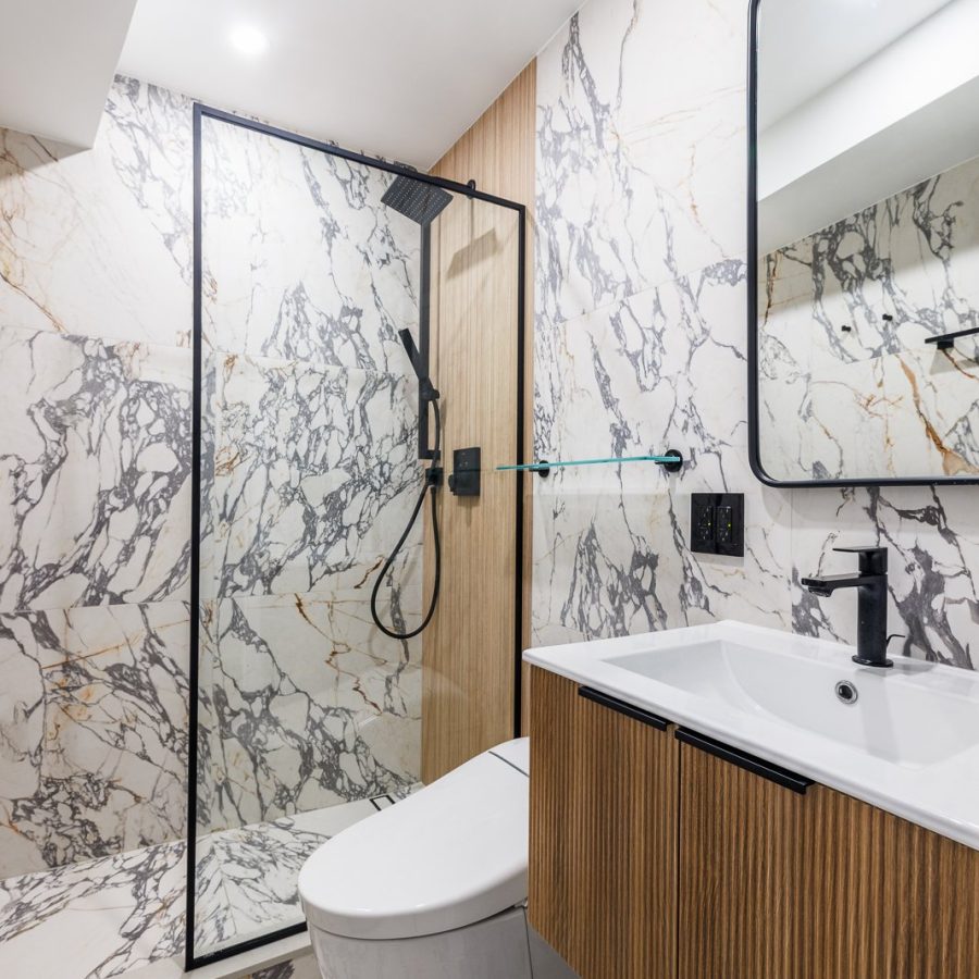 Contemporary bathroom with striking marble-look walls and flooring accented by black veining. The design features a frameless glass shower, a floating wood vanity with a white countertop, and a sleek rectangular mirror. Black fixtures and warm wood accents create a sophisticated contrast, blending modern minimalism with natural textures. Bathroom remodeling in Manhattan.