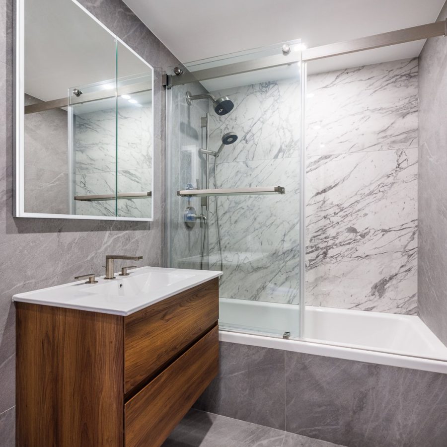 04932 Contemporary bathroom remodel featuring a floating wood vanity, backlit mirror, and a glass-enclosed tub with marble wall tile and gray porcelain flooring.