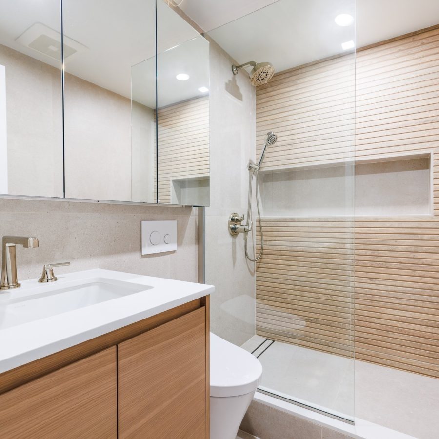 09347 Home remodeling contractor on the West Side. Modern bathroom with light wood vanity, minimalist fixtures, wall-mounted toilet, and a walk-in shower featuring horizontal wood-textured tile and glass enclosure.