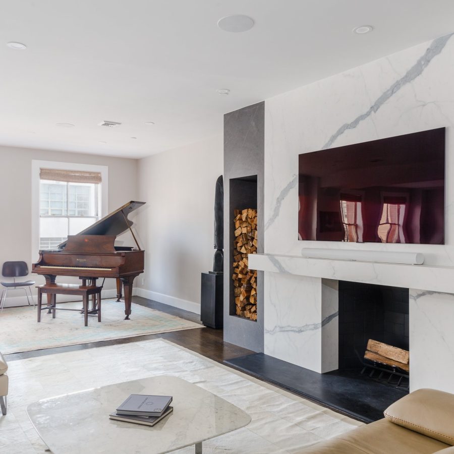 10114 Elegant living room featuring a sleek marble fireplace with a mounted flat-screen TV and built-in wood storage niche. The space includes a grand piano on a light area rug near a window, with natural light highlighting the clean lines and minimalist décor. A marble coffee table and neutral furnishings add to the modern yet warm atmosphere.