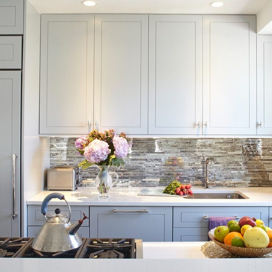 10208 Apartment renovation in Manhattan. Bright modern kitchen with light gray cabinets, a stainless steel backsplash, fresh flowers near the sink, a gas stove with a kettle, and a bowl of fruit on the island.
