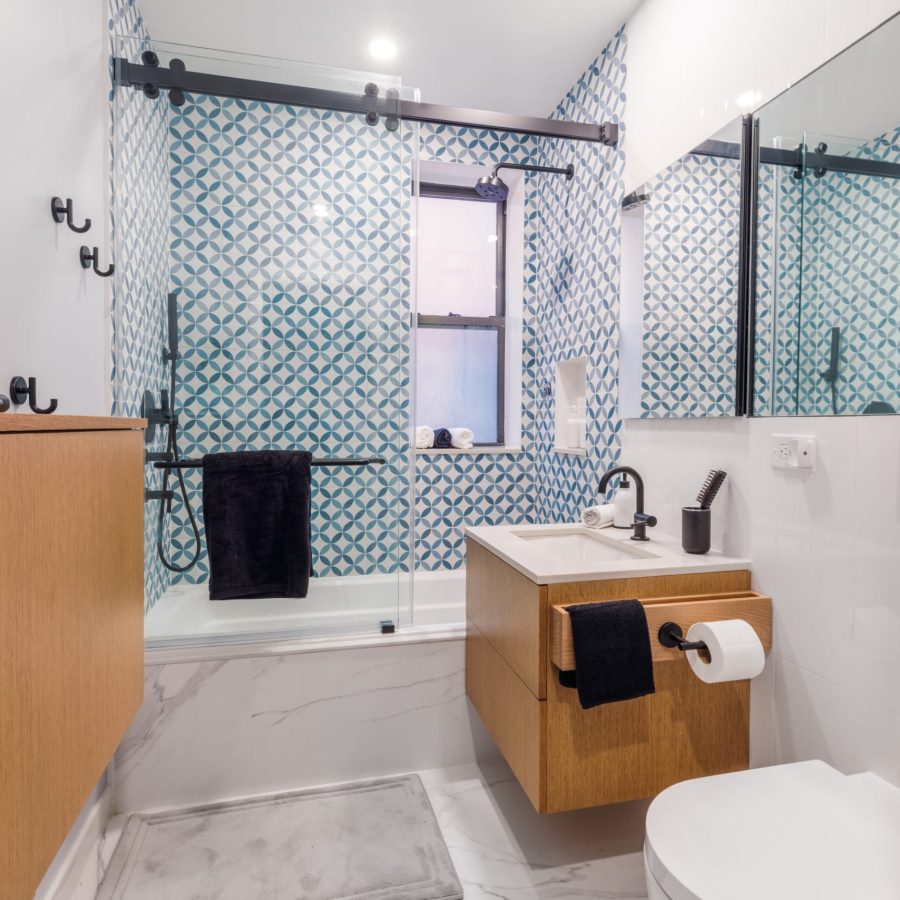 10332 Home remodeling contractor in Lincoln Square. Contemporary bathroom with a patterned blue and white tile shower wall, glass sliding shower doors, light wood vanity and cabinetry, wall-mounted sink, and modern black fixtures.