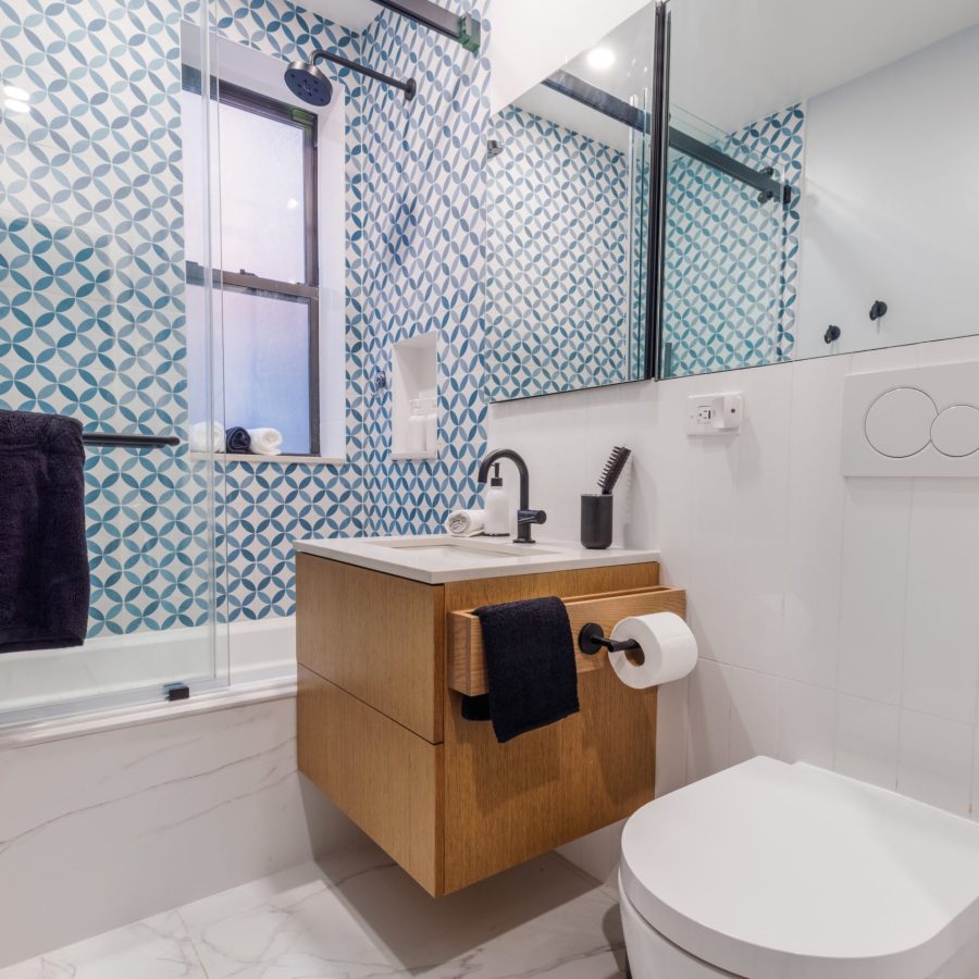 10337 Home remodeling contractor in the Upper East Side. Contemporary bathroom with patterned blue and white tile accent wall, floating wood vanity, black fixtures, glass shower enclosure, and white marble-style flooring.