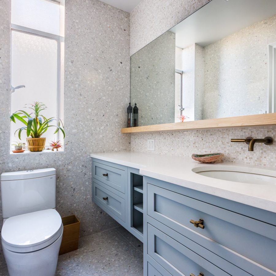 10341 Apartment renovation in Manhattan. Modern bathroom with terrazzo walls and flooring, a white toilet, light blue vanity with brass hardware, a large mirror, and a narrow window with potted plants.